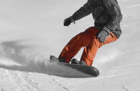 A snowboarder in a grey jacket and orange pants carving in the snow