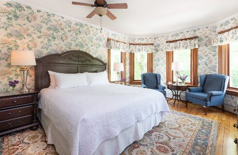 Spacious bedroom with king bed, two wingback chairs by four large windows and floral wallpaper