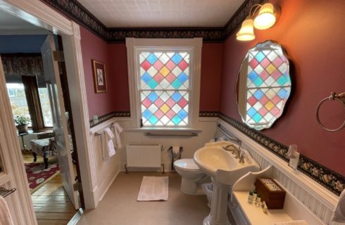 A bathroom with pedestal sink, toilet, oval mirror and large stained glass window