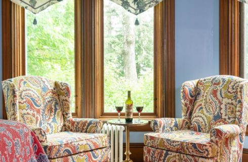 Two colorful wingback sitting chairs with table holding bottle of wine with two glasses in front of a window