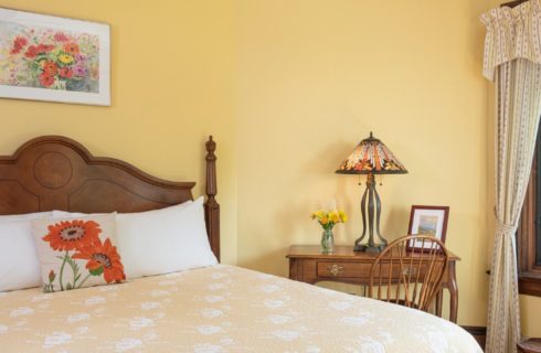 Bedroom with king bed with a yellow quilt, yellow walls, sitting table with lamp and chair by a window