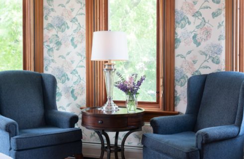 Two blue wingback chairs with round table holding a lamp and flowers by large bright windows