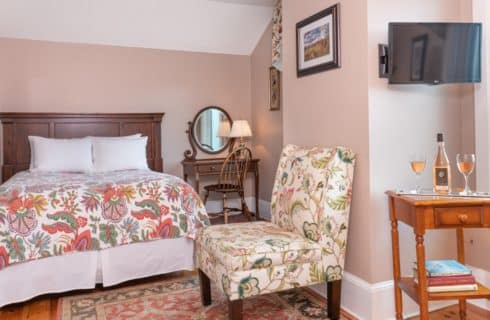 Small bedroom with a queen bed, floral quilt, dresser with oval mirror and chair, TV and table with wine glasses and bottle of wine