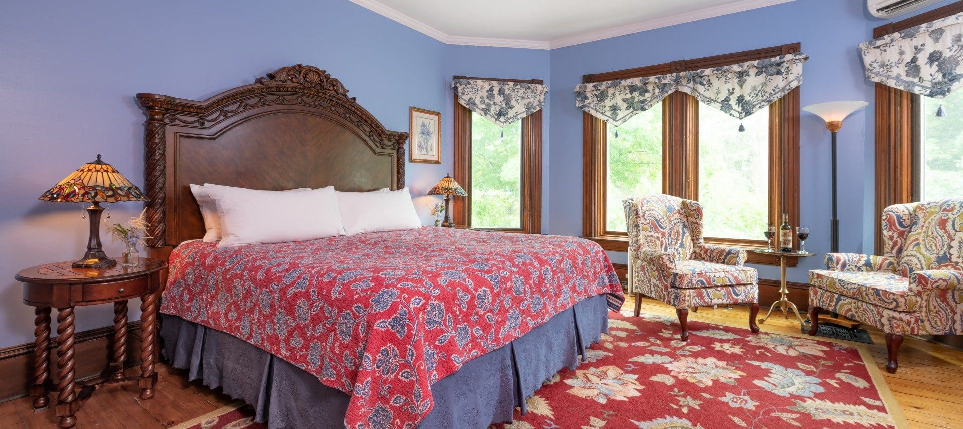 Large bedroom with a king bed in red and blue linens, four large windows with valence curtains and two wingback sitting chairs