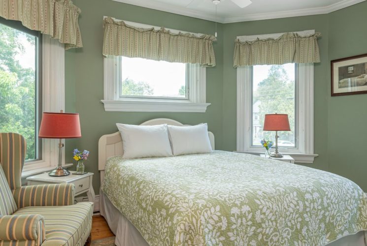 Pretty bedroom in green and white color palette with queen bed, striped chair and large windows, one with stained glass