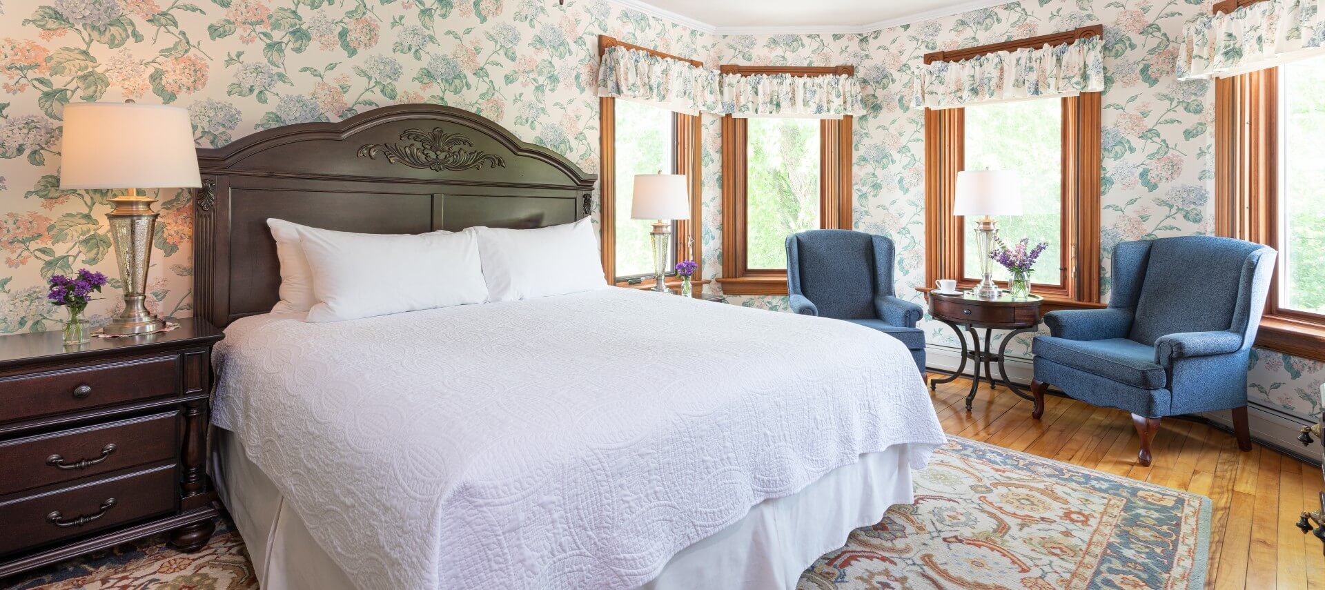 Large bedroom with king bed in white, blue wingback chairs by large windows and floral wallpaper and valences