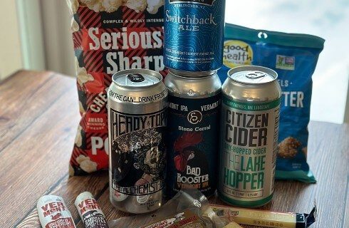 Several cans of different kinds of beer with two bags of snacks, meat sticks and cheese sticks